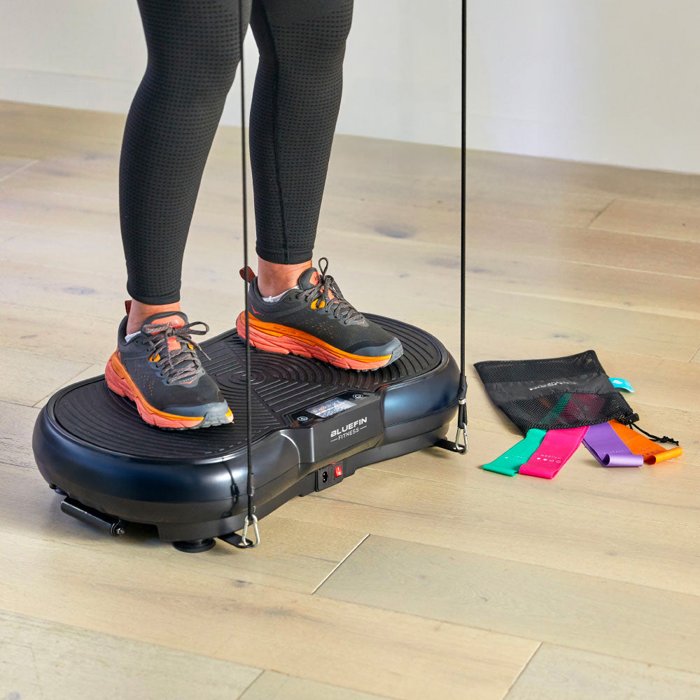 Person standing on a Bluefin fitness vibration plate with resistance bands, colorful exercise bands nearby.