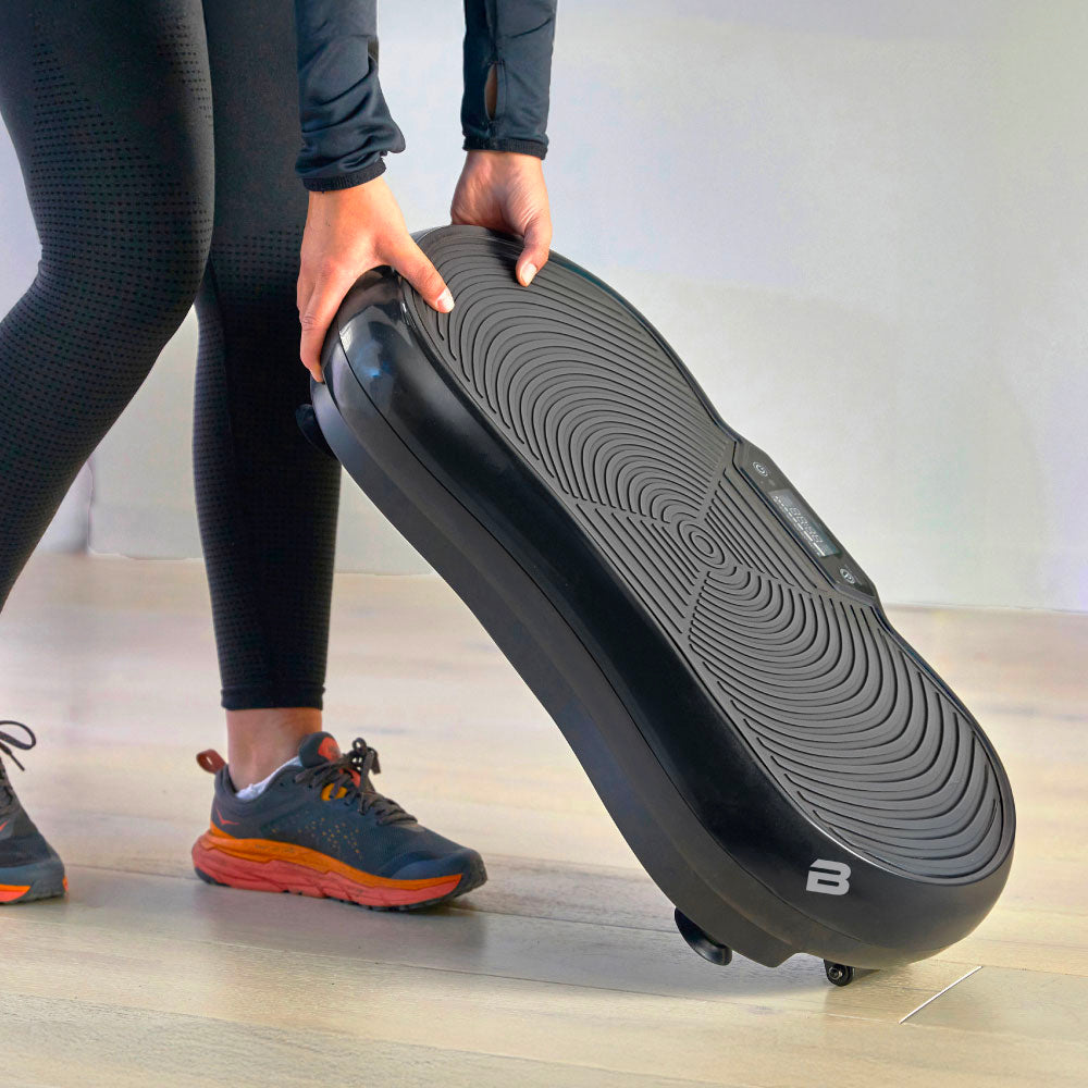 Person lifting a black fitness device with a textured surface and wheels, wearing black leggings and sneakers.