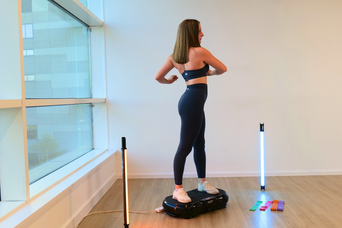 Woman standing on a vibration plate in a bright room, wearing black workout clothes and sneakers