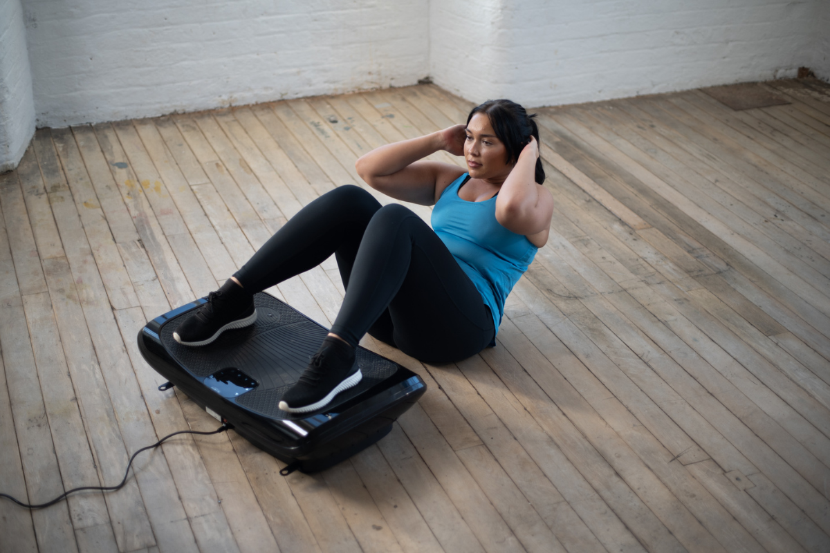 Woman performing abdominal crunches with her feet on a vibration plate in a room with wooden floors