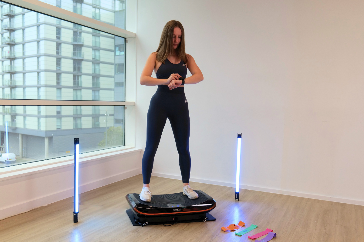 Woman standing on a vibration plate in a bright room, looking at her watch, with resistance bands on the floor beside her